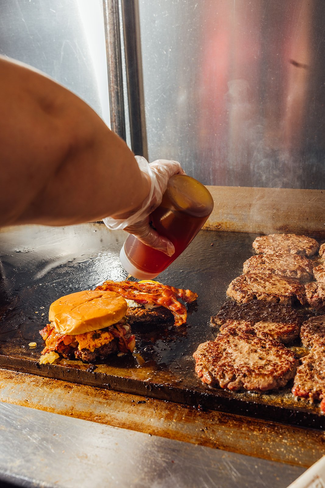Grillmaster adds sauce to a burger patty with toppings on a grill.