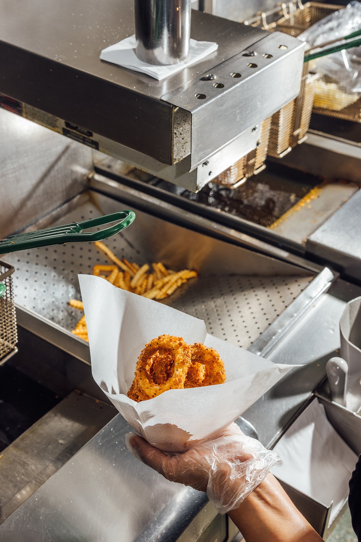 Person holding a paper basket with fried onion rings, in a food service kitchen with a fryer and cooking equipment in the background.