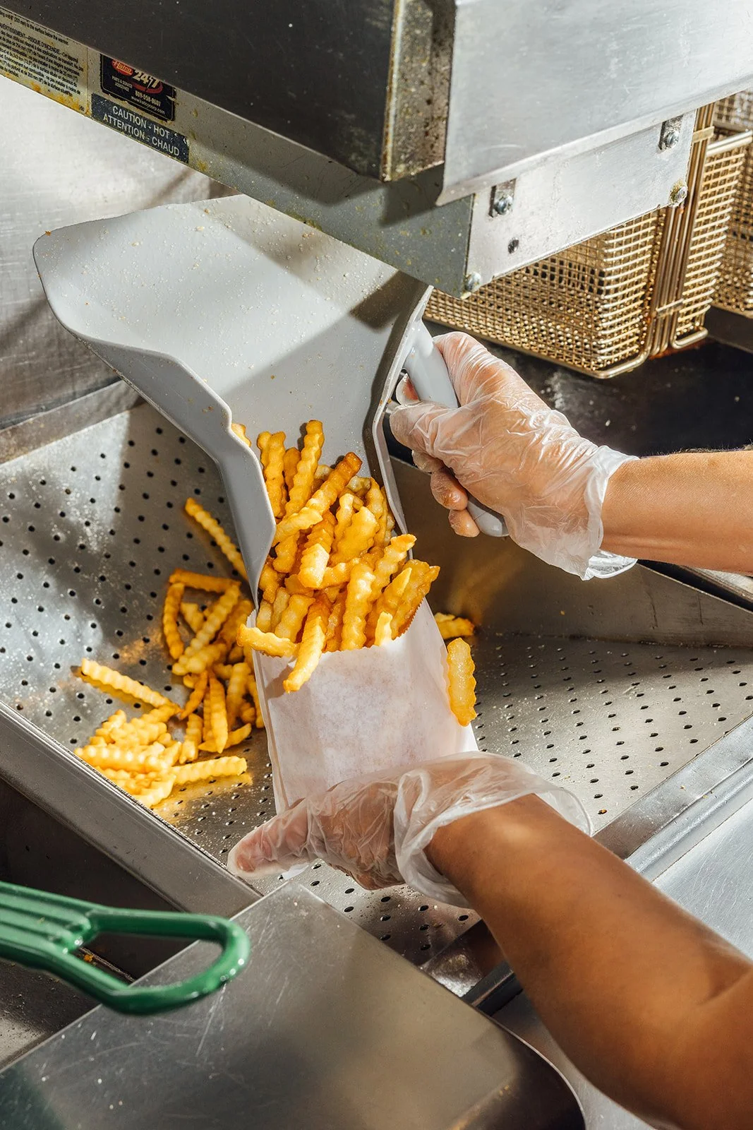 Person scooping fresh crinkle-cut French fries from a fryer into a paper bag.