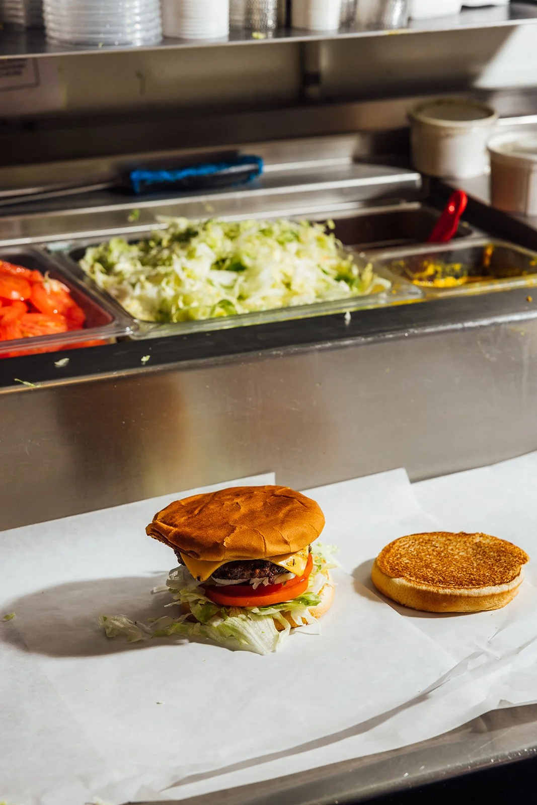 Unassembled cheeseburger with lettuce, tomato, cheeseburger patty, and bun on white parchment paper, next to a toasted bun in a fast-food kitchen with salad ingredients in the background.