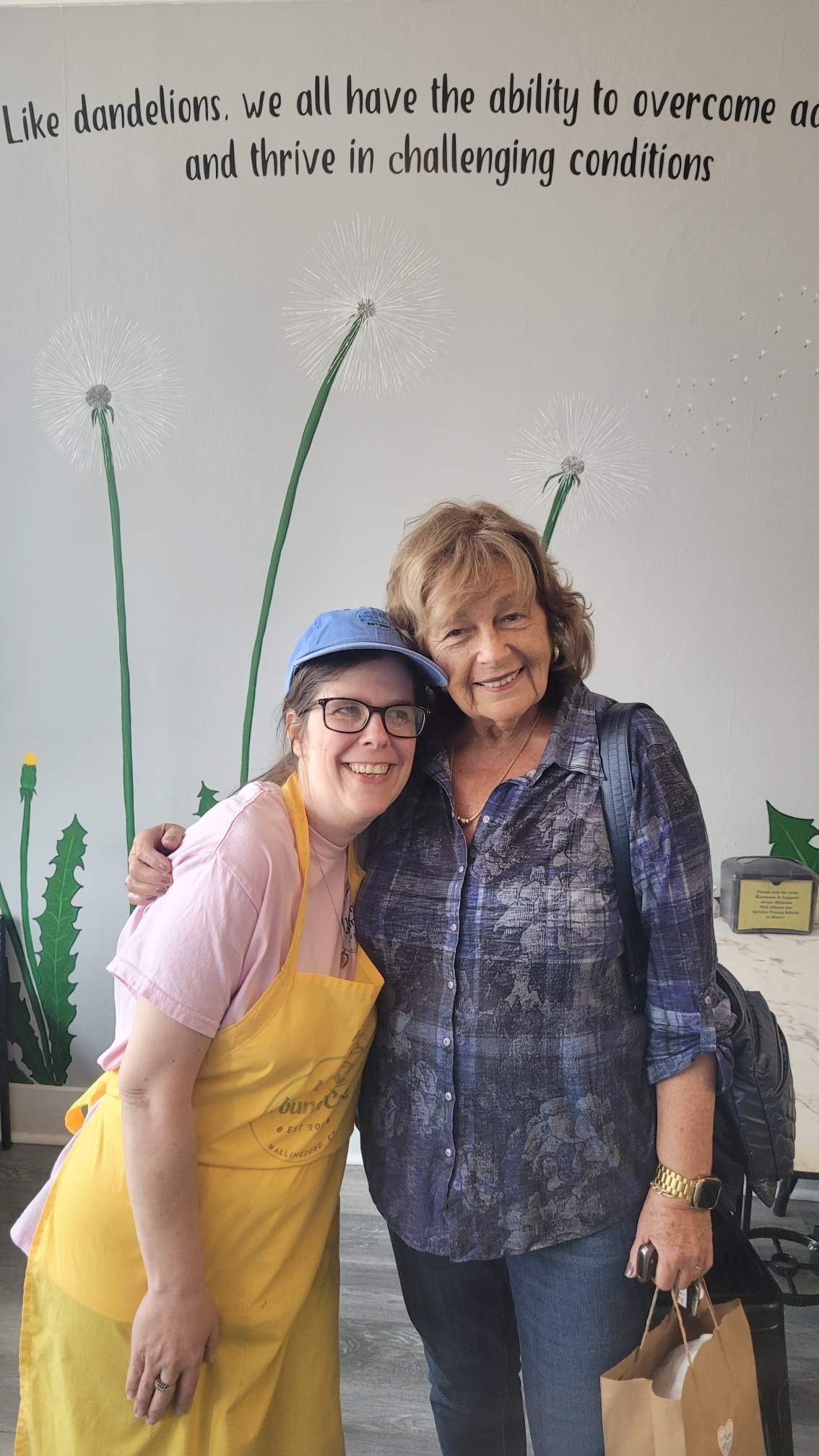 Two women are smiling and hugging in a room with a mural of dandelions on the wall behind them. One woman is wearing a yellow apron and a blue hat, while the other is wearing a blue patterned shirt and has a backpack.