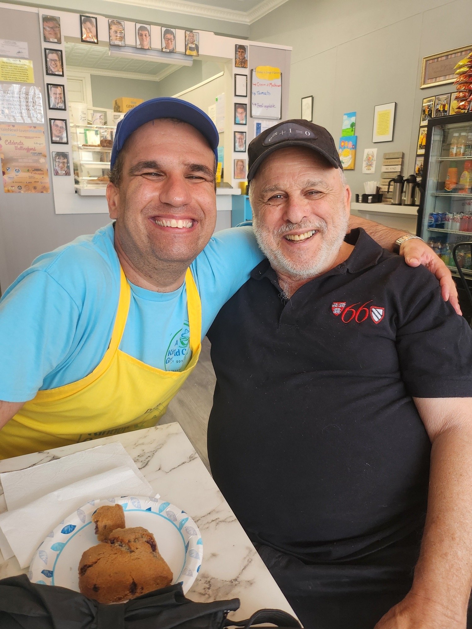 Two smiling men with their arms around each other sitting at a table in a cafe, with chocolate chip cookies on a paper plate in front of them. The man on the left is wearing a blue cap, a light blue shirt, and a yellow apron, while the man on the rig