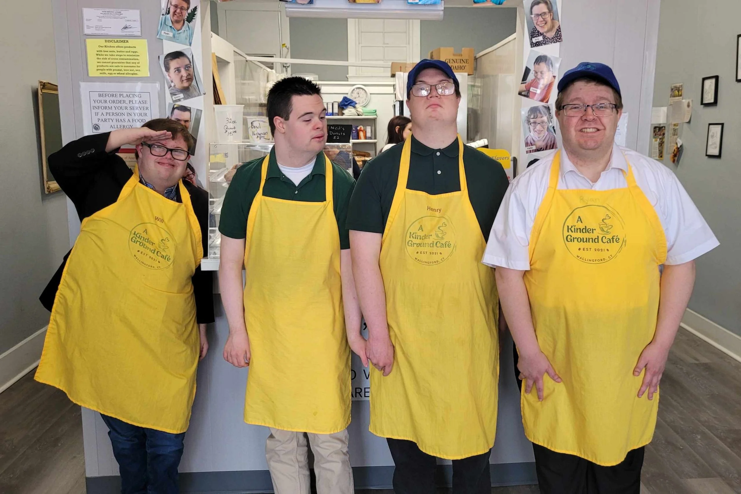 Four male staff members at the Kinder Ground Café, wearing yellow aprons and standing in line behind the counter. The background shows a countertop, some signs, photos of smiling people, and various cafe items.
