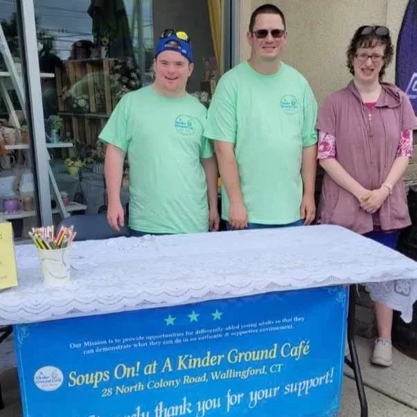 Three people standing behind a table with a blue banner, part of a fundraiser event at a kinder ground cafe, with the banner reading "Soups On! at A Kinder Ground Cafe" and the address 28 North Colony Road, Wallingford, CT. They are outdoors, smiling