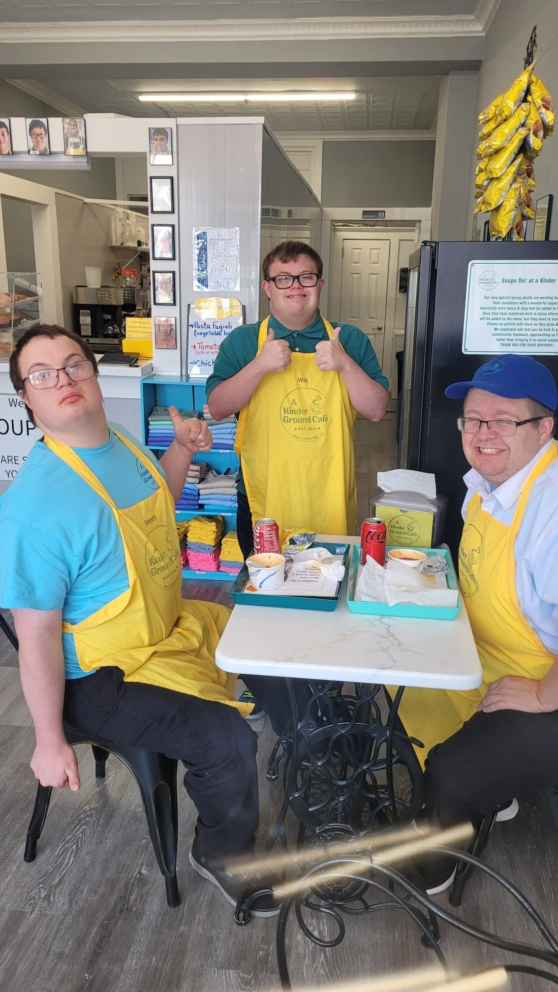 Three men wearing yellow aprons and glasses sitting and standing behind a white table with food and drinks in a cafe, smiling.