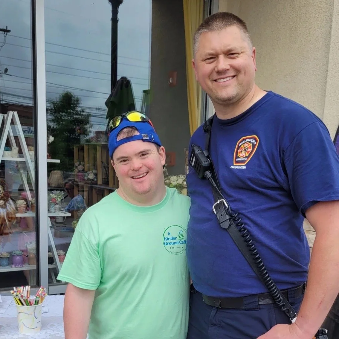 A young man with a blue baseball cap and green shirt standing next to a firefighter in a dark blue uniform outside a storefront with glass windows.