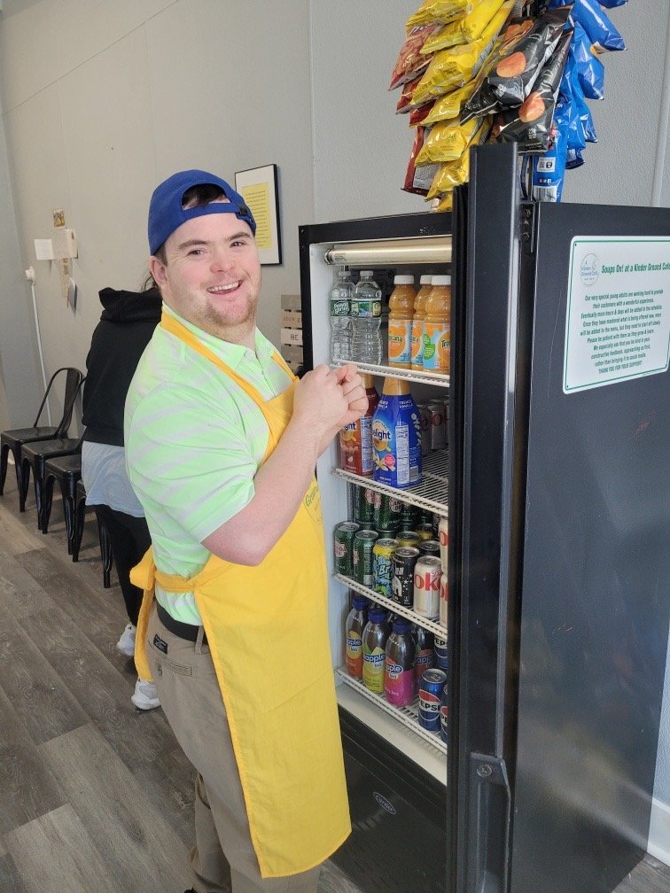 A smiling man wearing a blue cap, light green shirt, and yellow apron stands next to a refrigerator filled with bottled beverages. Items on top of the fridge include snack bags hanging in a rack. The background shows a few black chairs and a wall wit