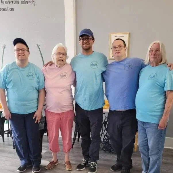 Group of five people standing side by side in a room, wearing matching blue and pink t-shirts, smiling at the camera.