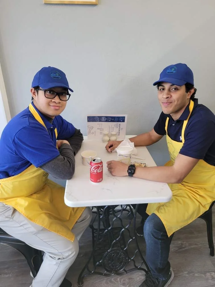Two men sitting at a small white table in a casual eatery, wearing blue caps and yellow aprons. One man has glasses, and the other is smiling. On the table are small cups, a red Coca-Cola can, and a menu or informational sheet.