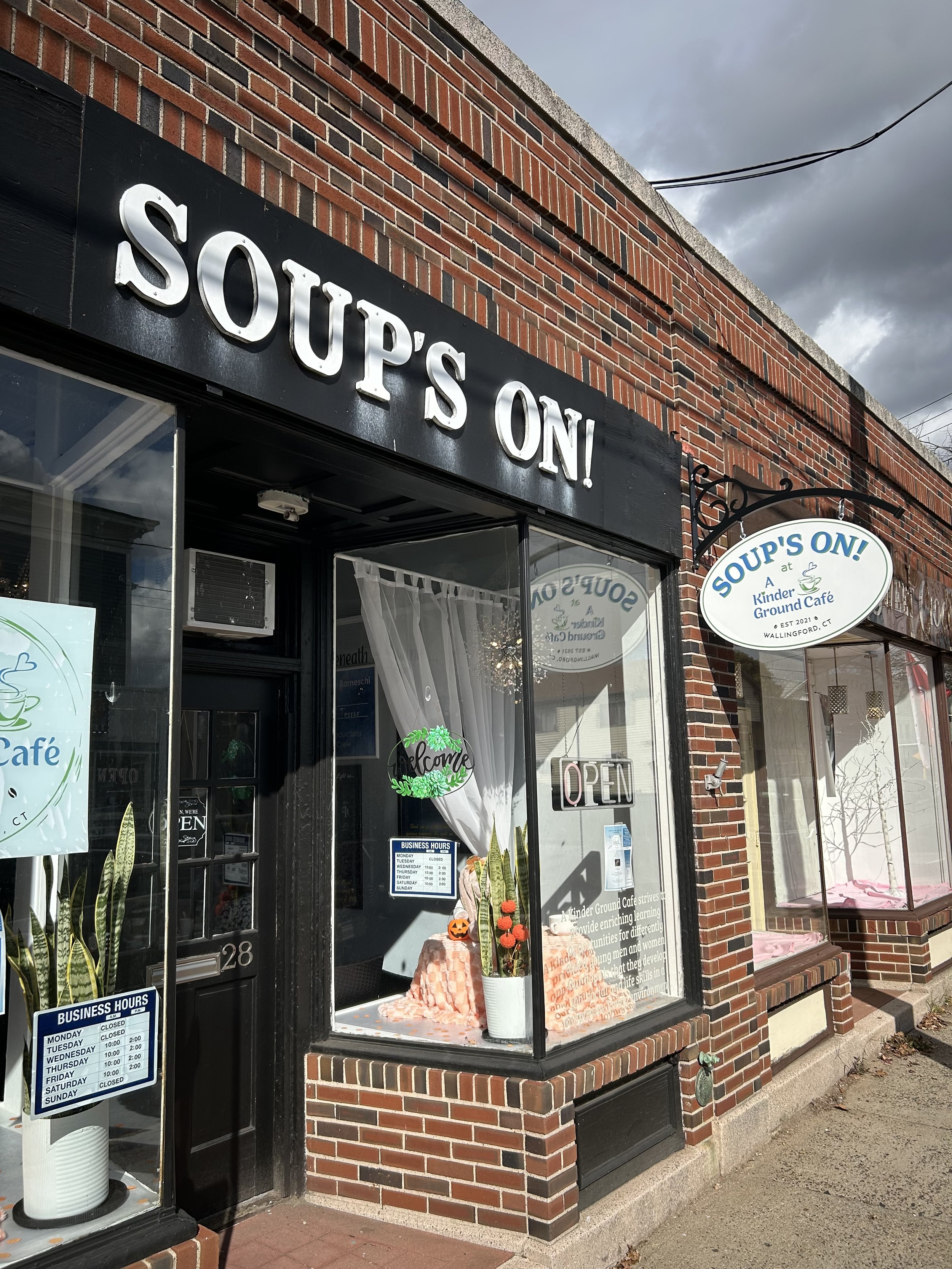 A storefront with black and white signage that reads 'SOUPS ON!' and a smaller round sign that says 'SOUPS ON!' The window display includes a sign with business hours, a potted cactus, and a 'Welcome' sign. The sidewalk in front is concrete with some leaves.