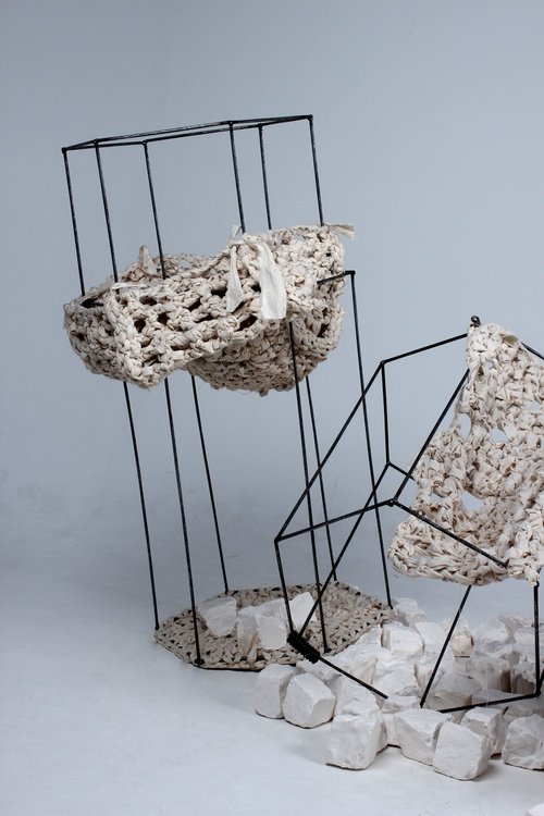 Art installation with wireframe chairs, one with a textured, irregular surface and the other with similar construction, surrounded by white rocks on a plain background.