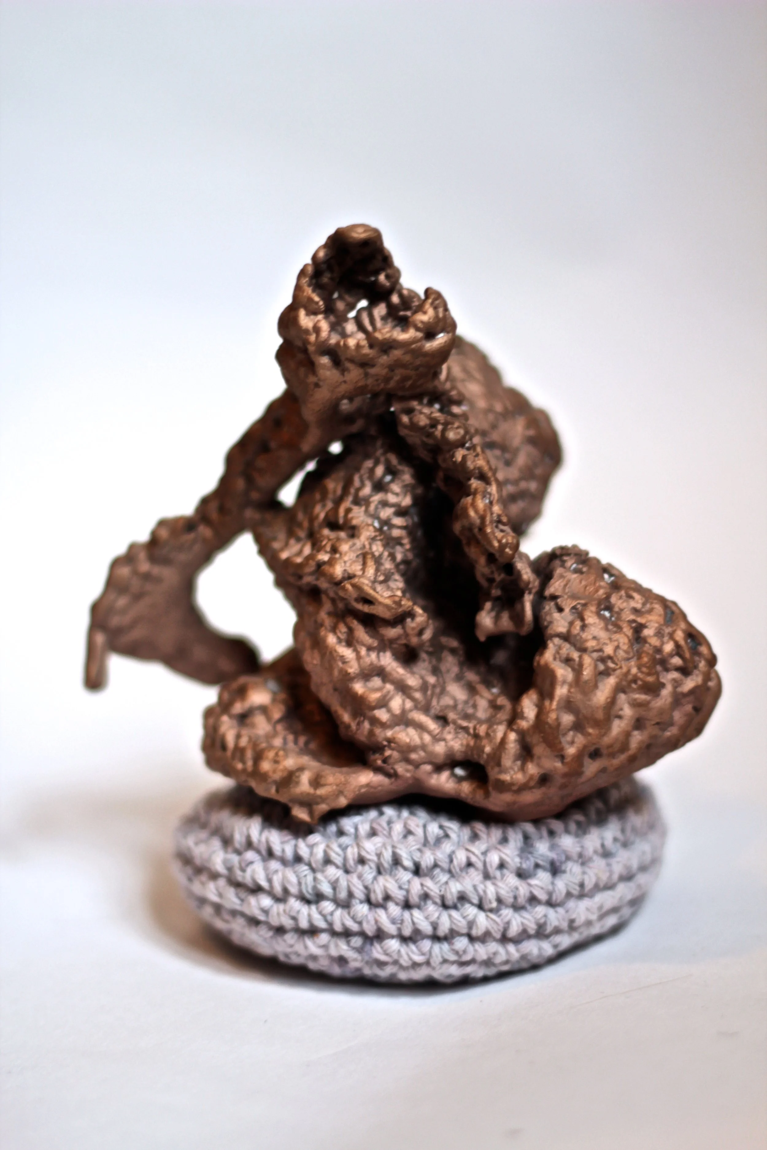 Close-up of a small sculpture of a praying mantis made from twisted brown wire, sitting on a crocheted gray base.