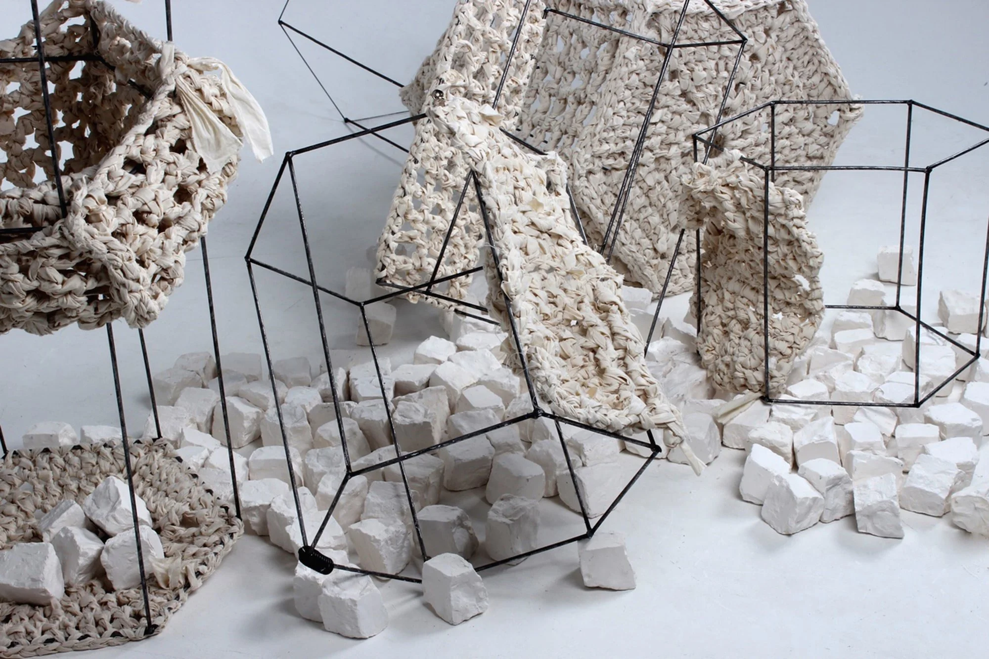 Art installation consisting of black wire frames holding large pieces of knotted white fabric, surrounded by white foam blocks on a white floor.