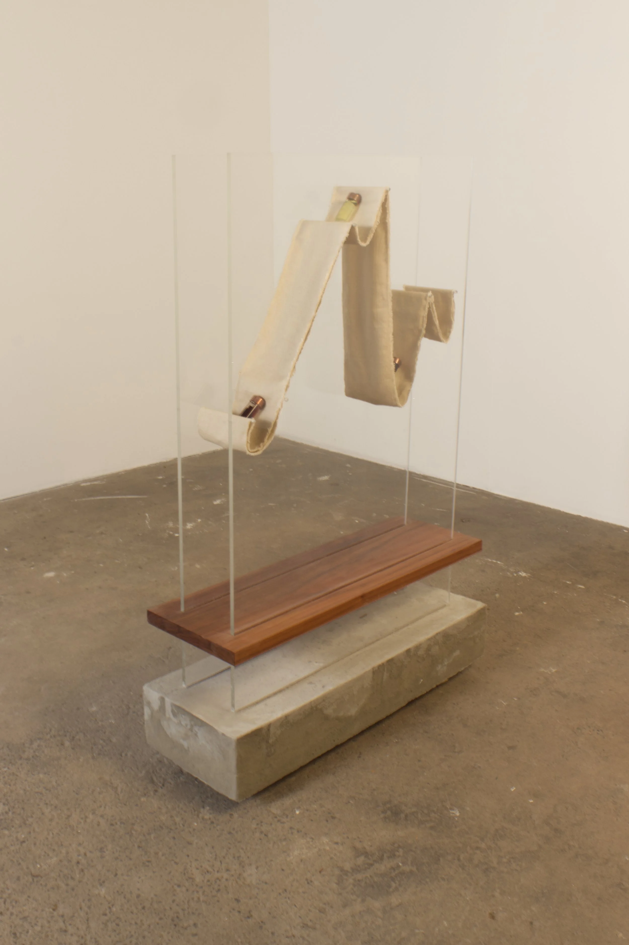 An art installation featuring two fabric strips, one beige and one light brown, draped over copper rods, displayed on a clear acrylic stand with a wooden base, set on a concrete floor.