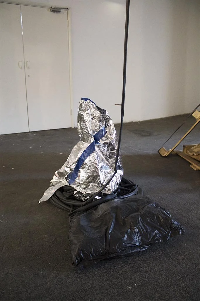 A pile of black plastic bags with a foil-covered object and a pole in a room with plain walls and a door.