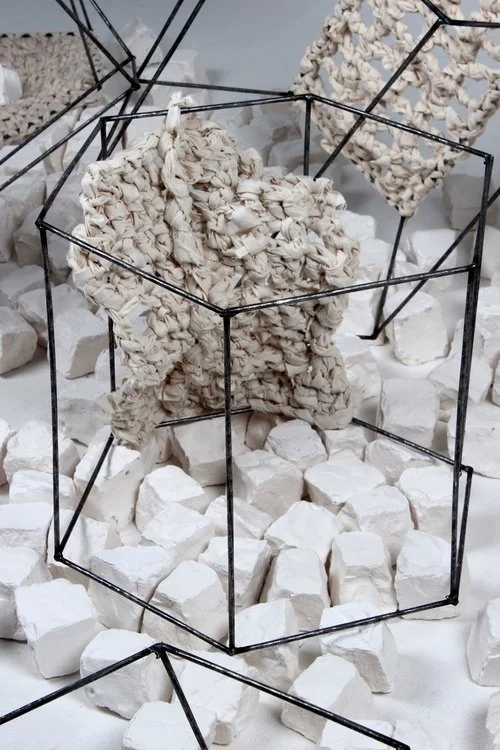 Abstract art installation with white stone-like blocks, black wireframes, and textured fabric or paper sculptures.