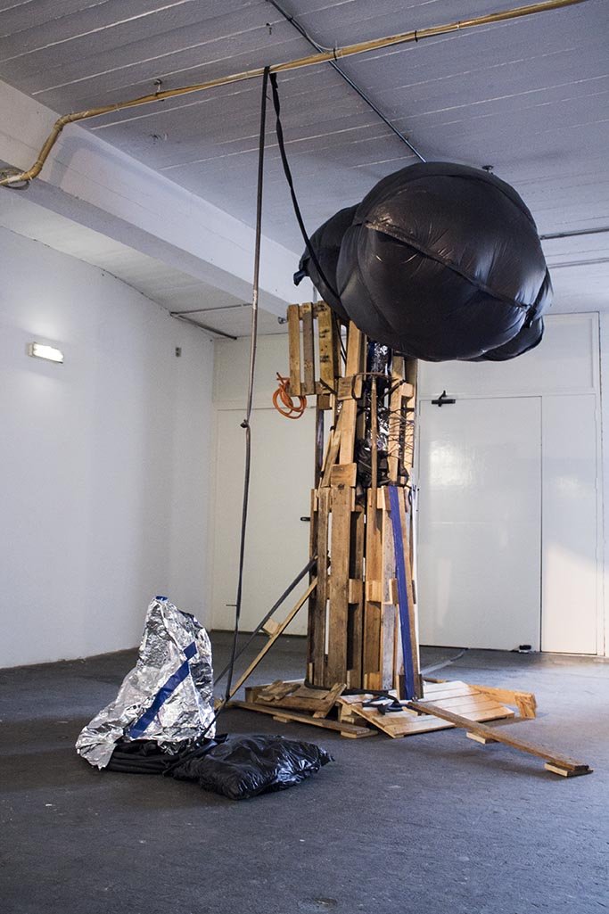 An art installation in a white-walled studio features a structure made from wooden pallets and planks, with a large black inflated object hanging from the ceiling, connected by black straps. Foil and black plastic bags are on the floor nearby.