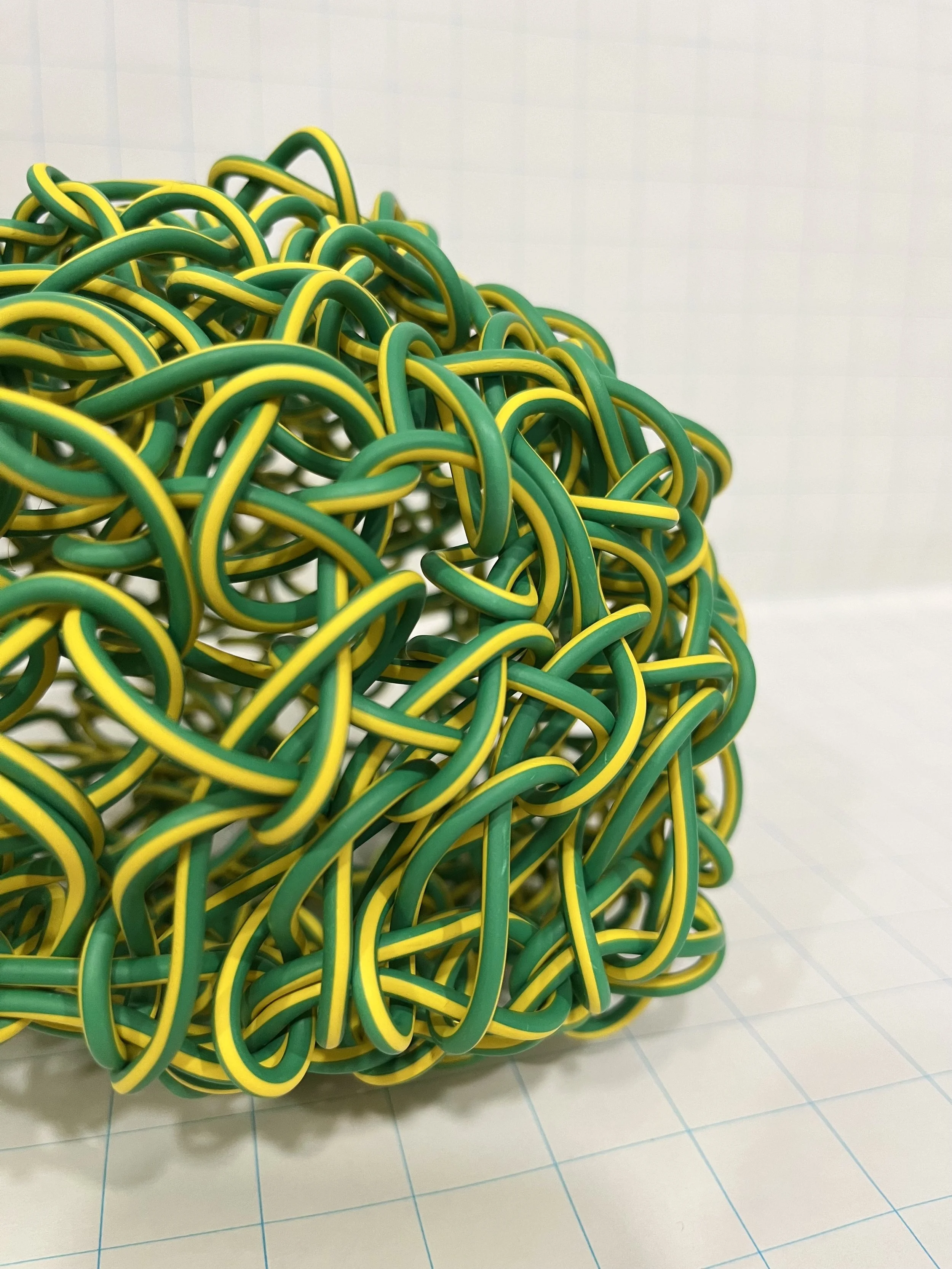 An art installation or sculpture made from intertwined green and yellow rubber bands or elastic. The structure is placed on a tiled floor with a grid pattern, against a plain wall background.