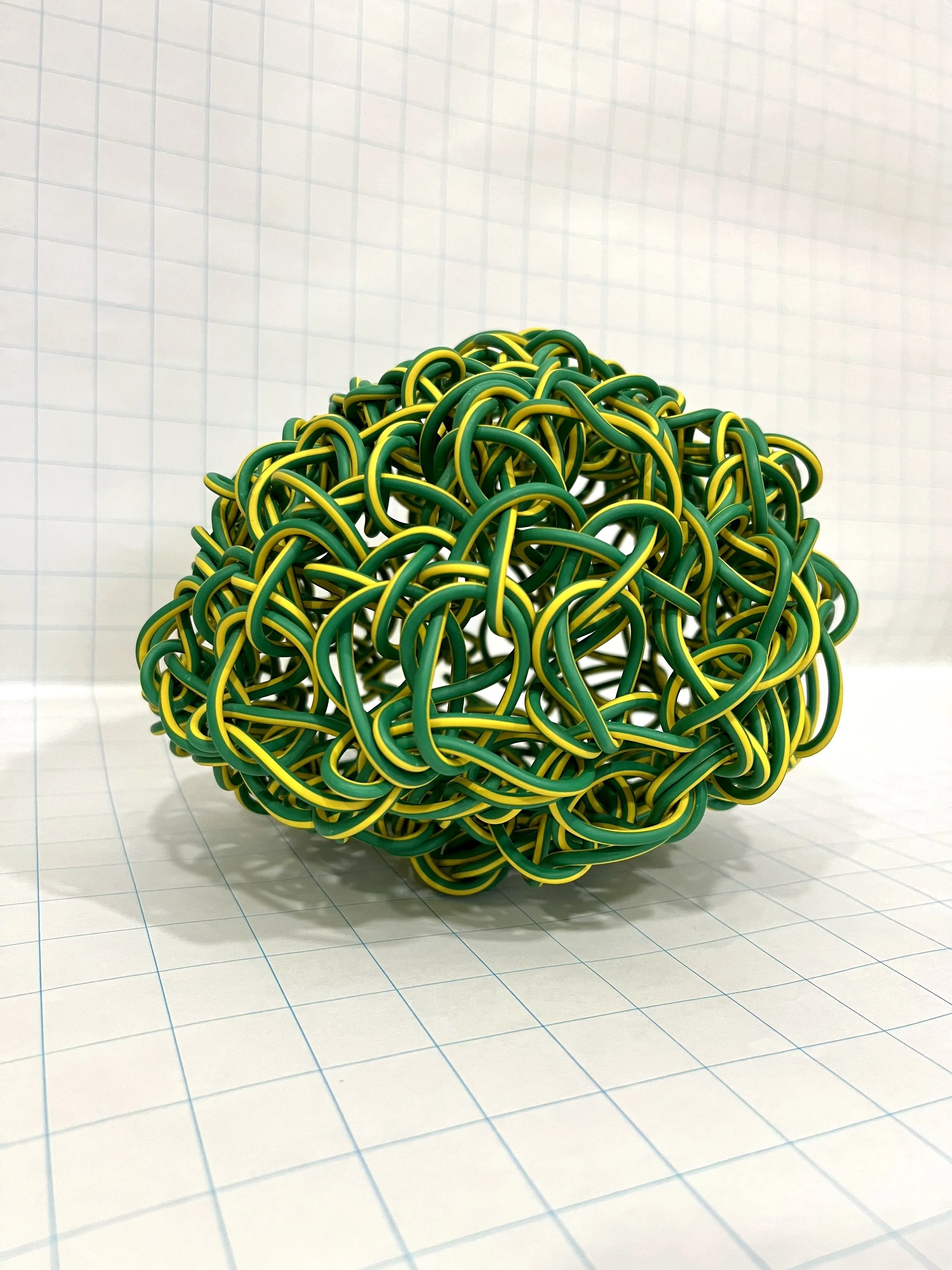 A colorful ball made of intertwined yellow and green rubber bands placed on a white grid paper surface with blue grid lines and a light background.