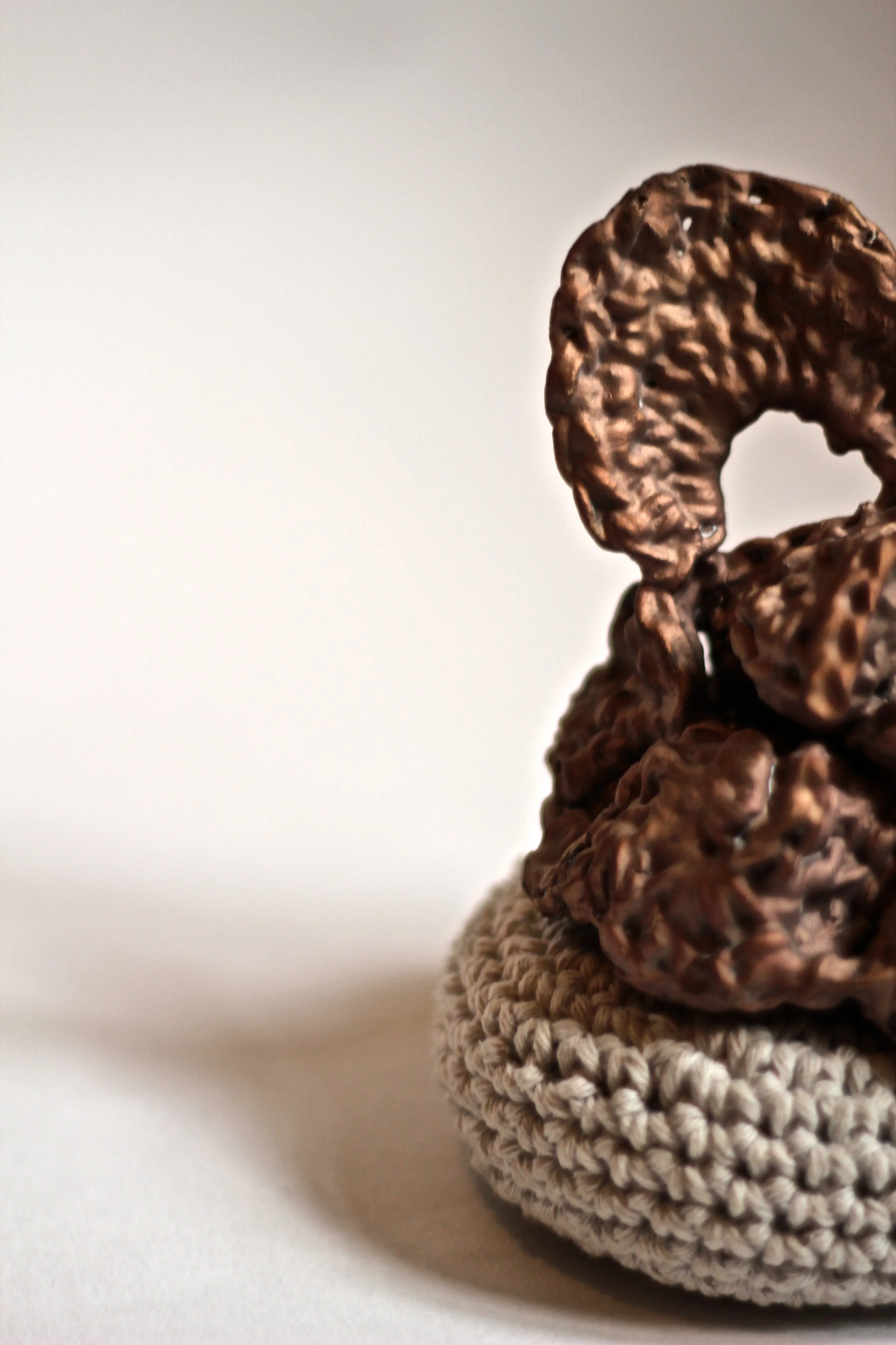 Close-up image of a crocheted beige hat with a large, decorative metal ring on top, set against a plain white background.