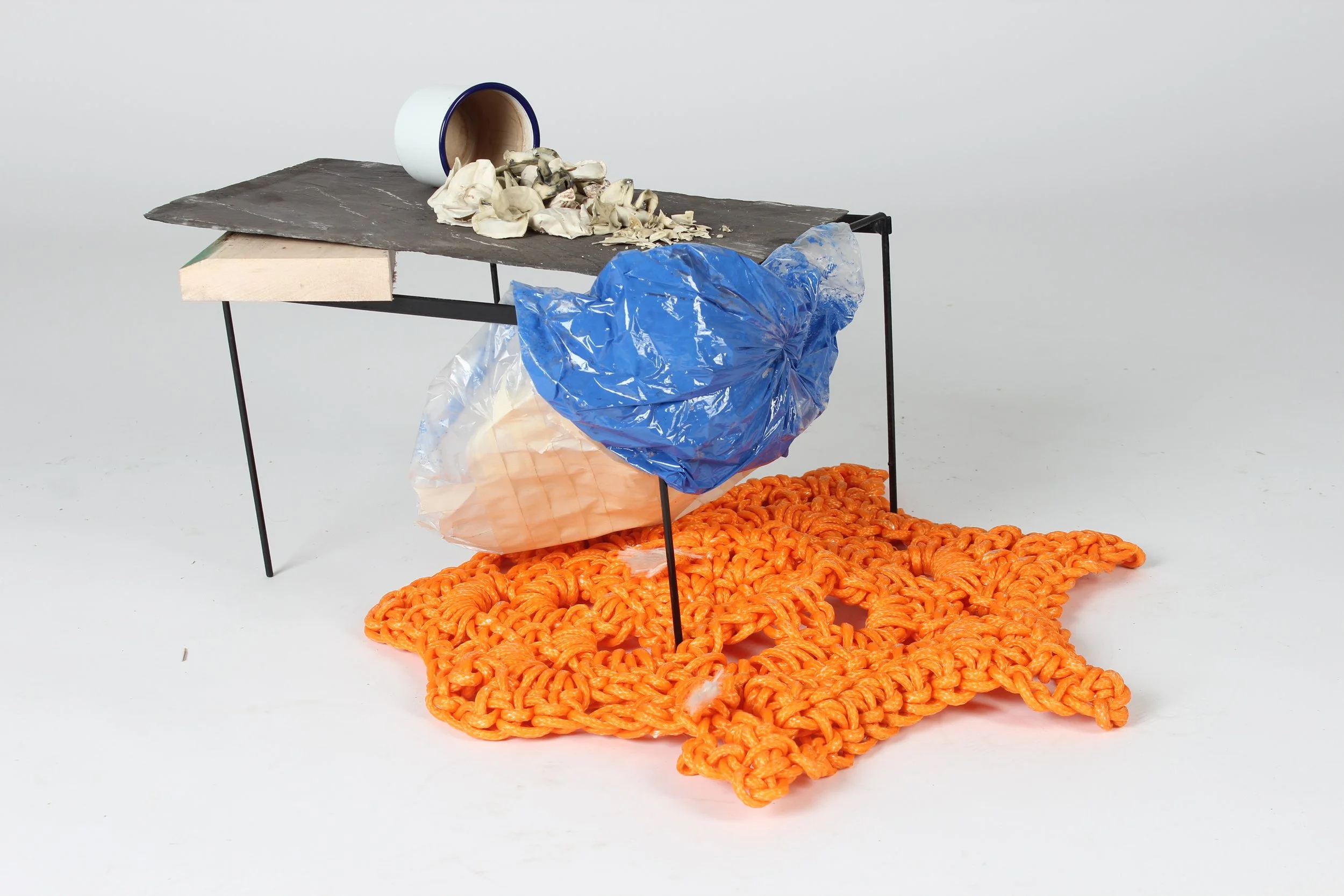A small table holds a broken white mug with a blue rim that is spilling crumpled paper, a large pile of orange plastic zip ties, and a crumpled blue plastic bag. The background is plain white.