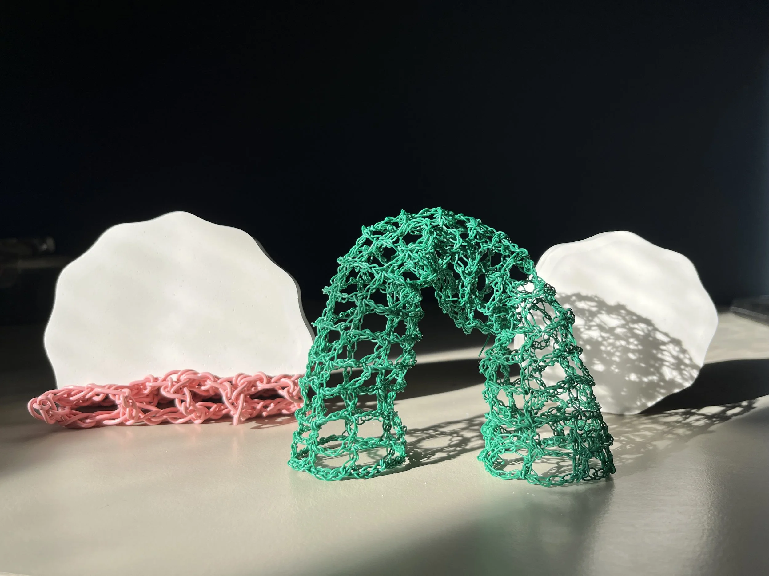 Various colorful woven and chain-like sculptures, including a green woven figure, a pink chain, and white abstract shapes, displayed on a white surface against a dark background.