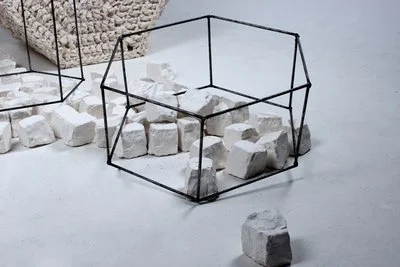 A geometric wireframe container with white stones inside and scattered around on a plain surface.