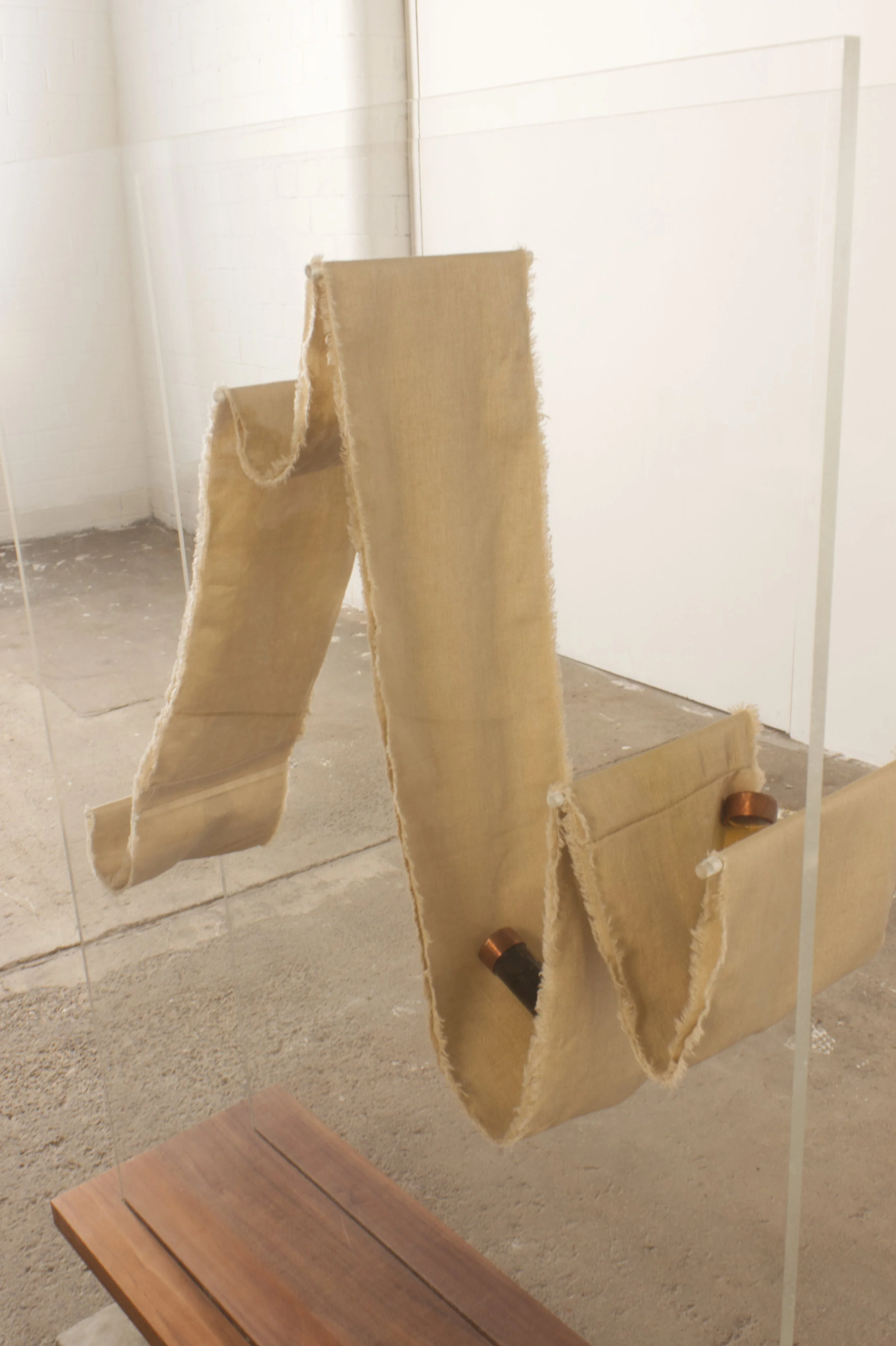 A mirror in an art gallery or museum reflects hanging beige fabric strips with frayed edges and copper rollers, in an industrial space with a white brick wall and concrete floor.