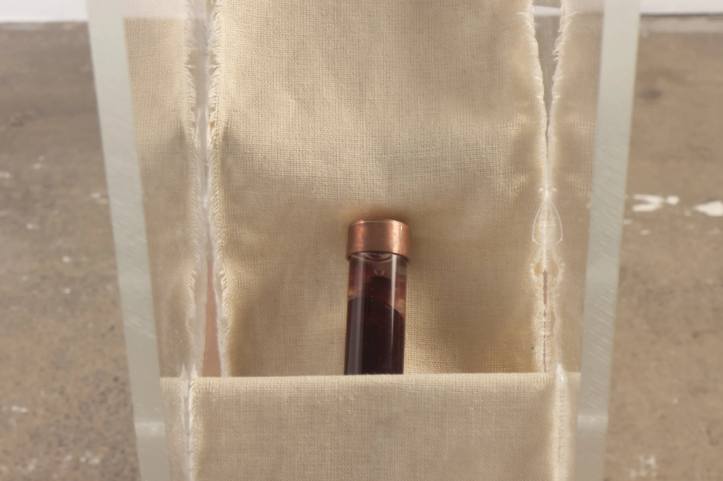 A glass test tube with dark liquid inside, standing on a beige fabric background with fringe edges.