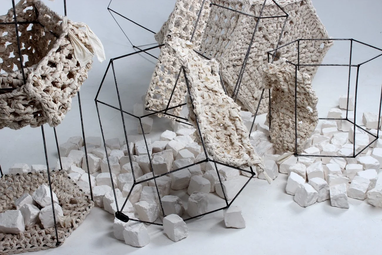 An art installation featuring multiple geometric wireframes and woven fabric pieces, with white carved rocks scattered on the floor.