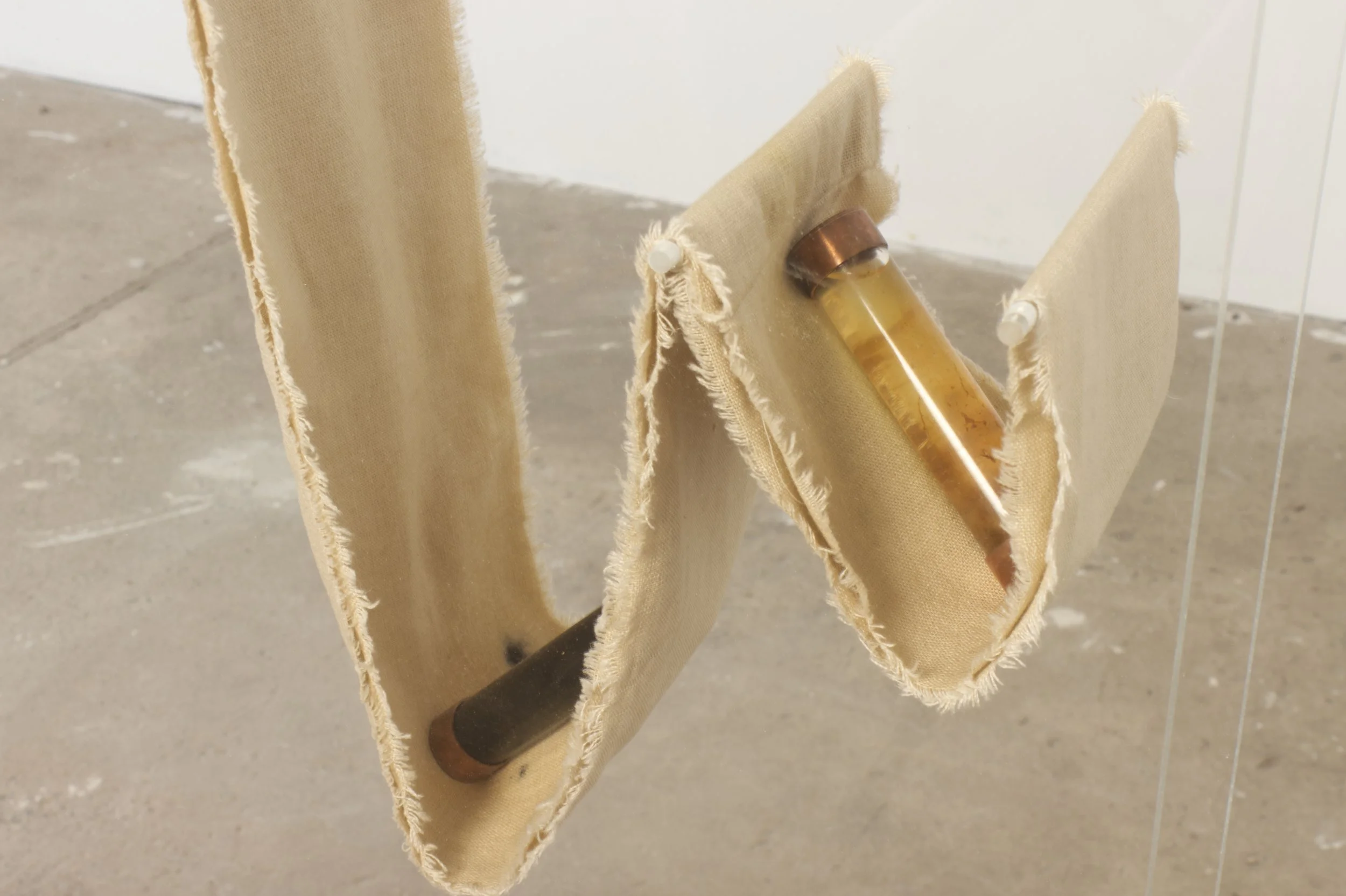 Close-up of four beige fabric pieces hanging on white pegs, with a glass vial containing a yellowish liquid leaning against one of the fabric pieces.