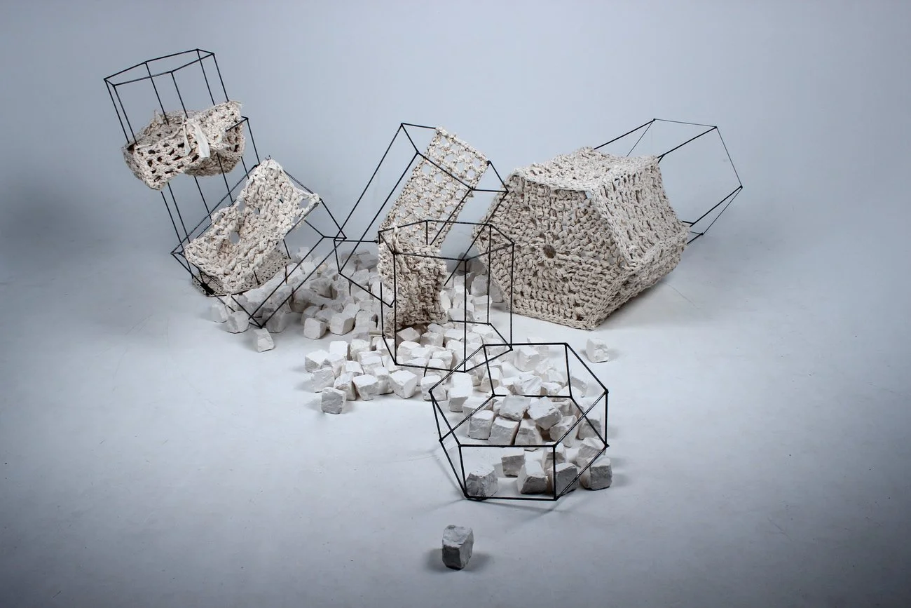 Contemporary art installation featuring broken white ceramic blocks, some held within black metal wireframe structures, with scattered pieces on a light gray surface.