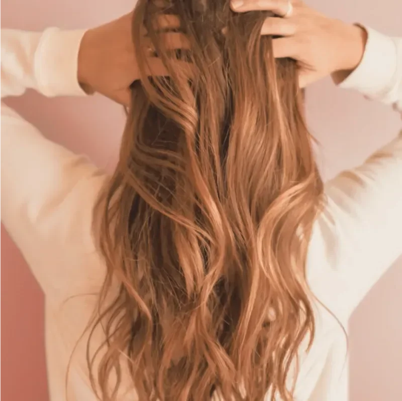 Back view of a woman with long, wavy, reddish-brown hair, wearing a white top, with hands in her hair against a pink background.