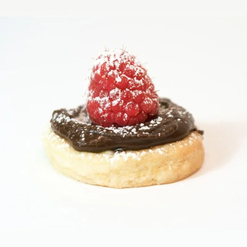 Close-up of a pastry topped with a chocolate spread, a raspberry dusted with powdered sugar, on a white background.