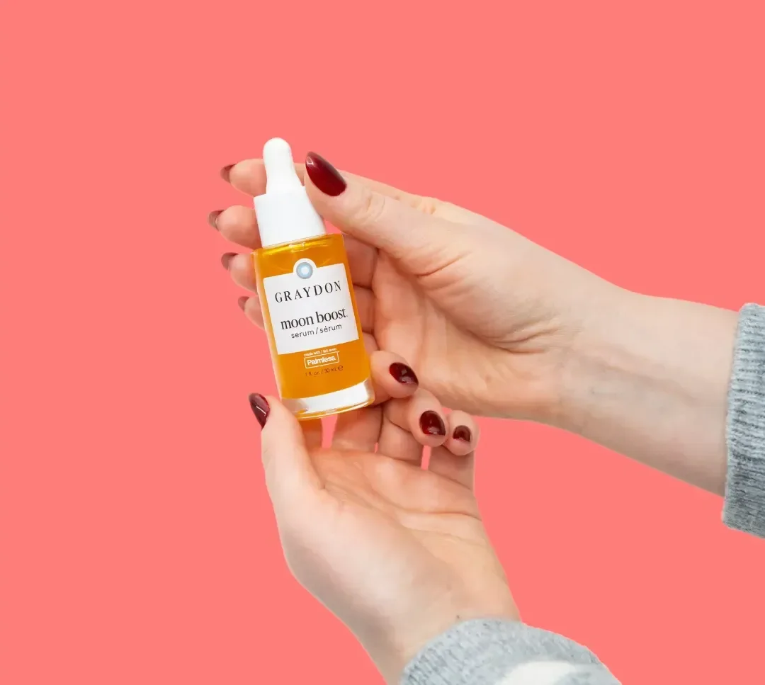A person's hands holding a small bottle of Graydon moon boost serum against a pink background.