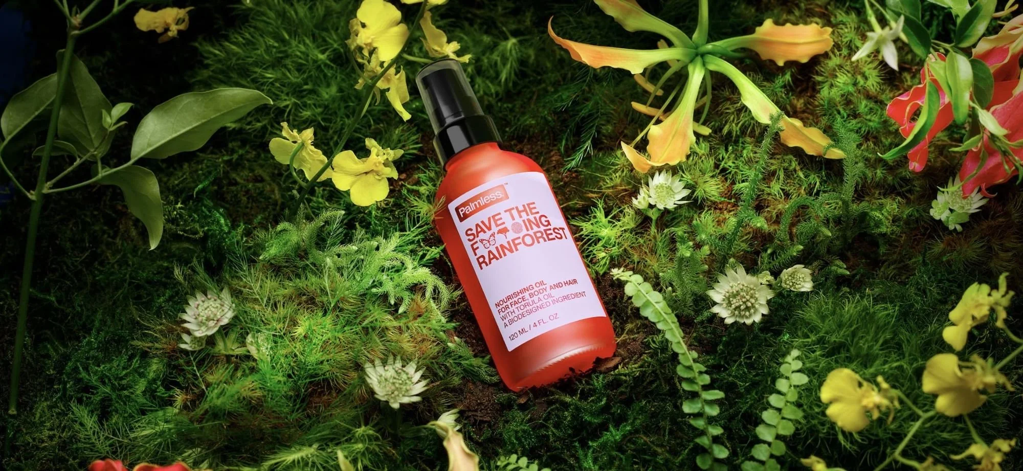 A bottle of Palmeless Save the Rainforest nourishing oil lying on green moss surrounded by various green and yellow plants and flowers.