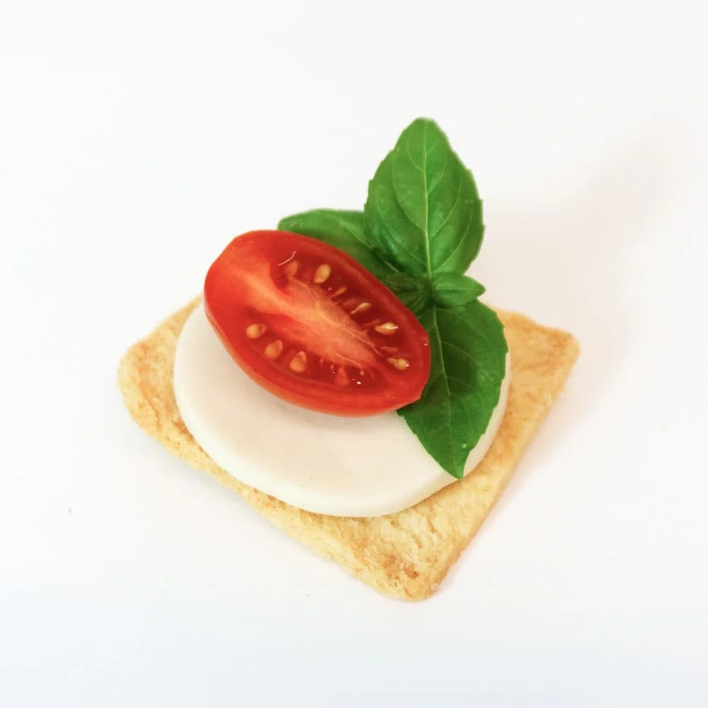 An open-faced snack with a square piece of bread, topped with a round slice of mozzarella cheese, garnished with a cherry tomato half and fresh basil leaves.