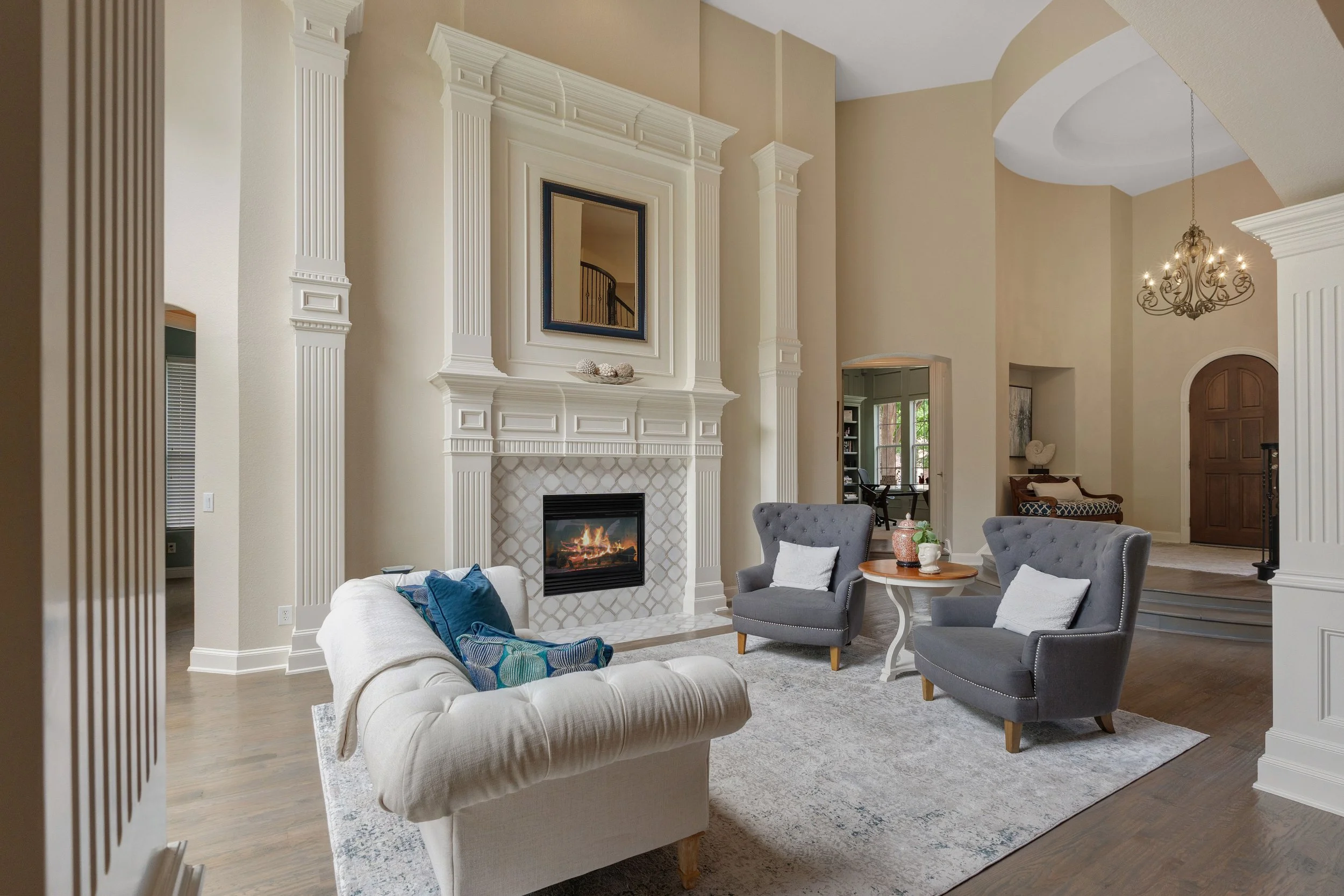 Living room with a stone fireplace, high ceiling, chandelier, beige walls, and a white sofa with blue and white pillows, gray armchairs, a round side table with decor, and a rug on hardwood floor.