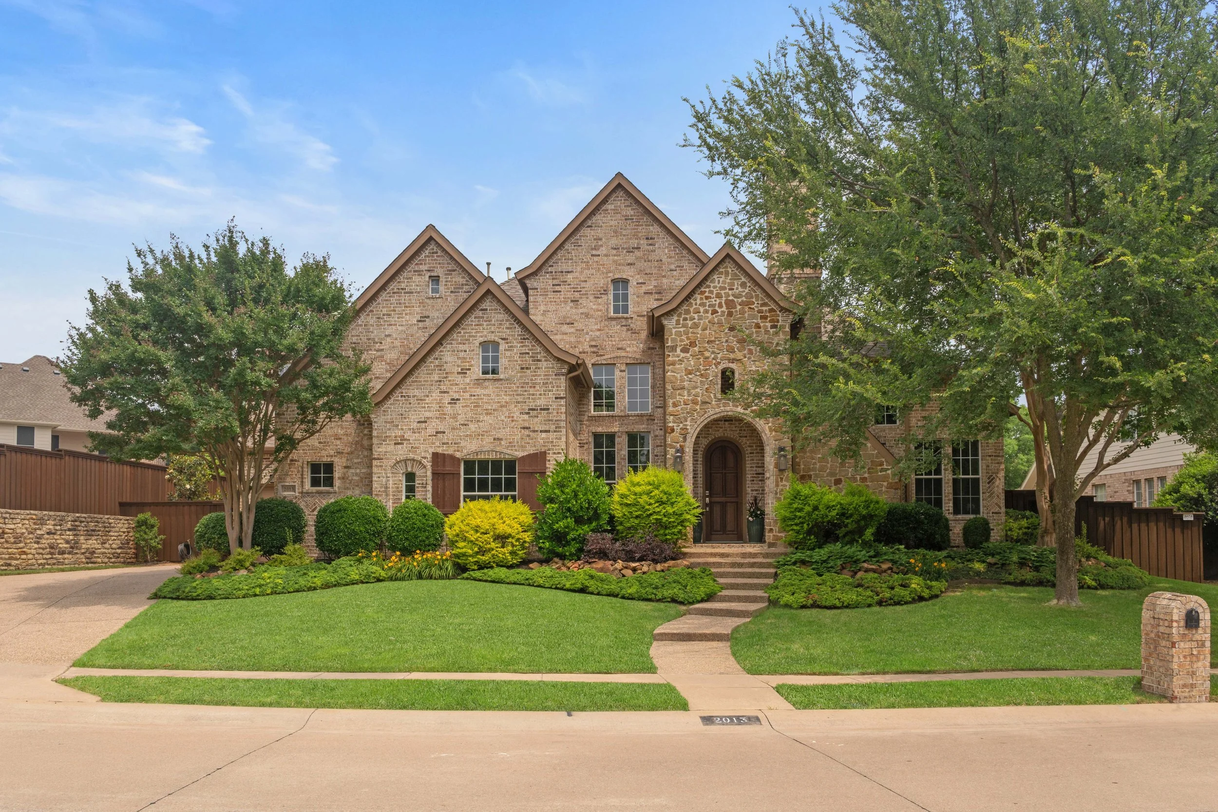 Front view of a large brick house with a manicured lawn and landscaping, including trees, bushes, and flower beds, under a blue sky.