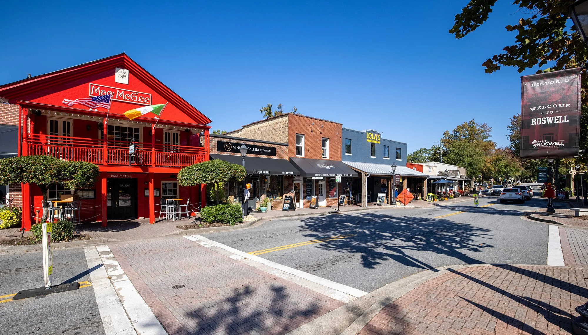 A street scene in Roswell featuring a bright red building with a balcony, a row of shops including an art gallery and a restaurant, trees, and cars parked along the road on a sunny day.