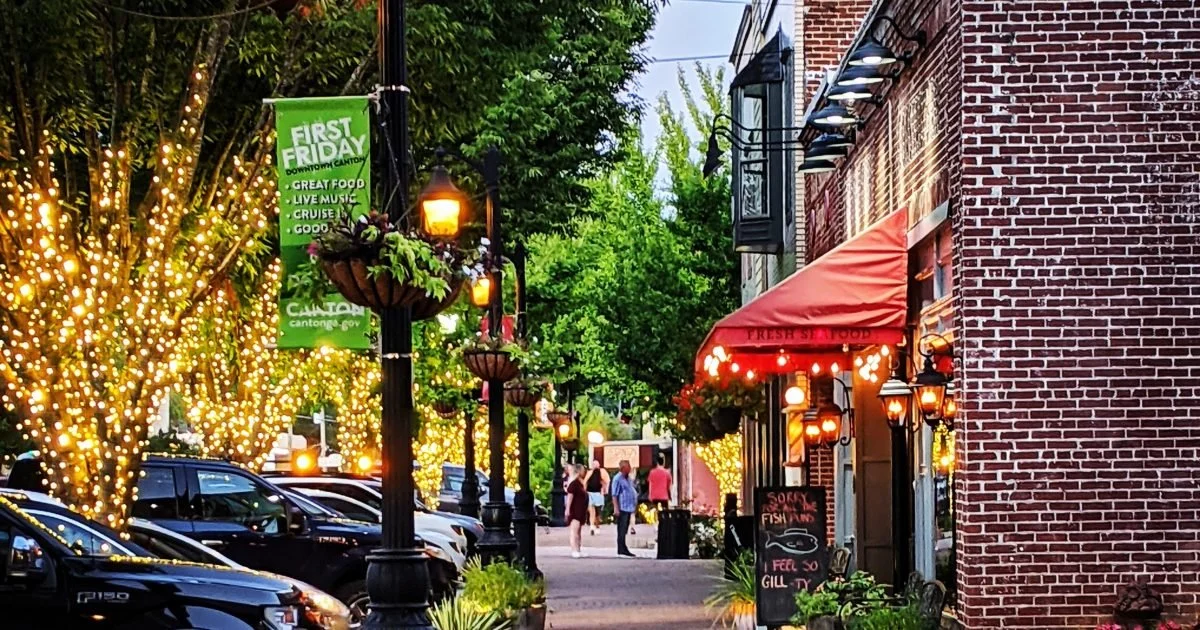 A lively downtown street with trees wrapped in string lights, parked cars, and a restaurant with a red awning and hanging lanterns.