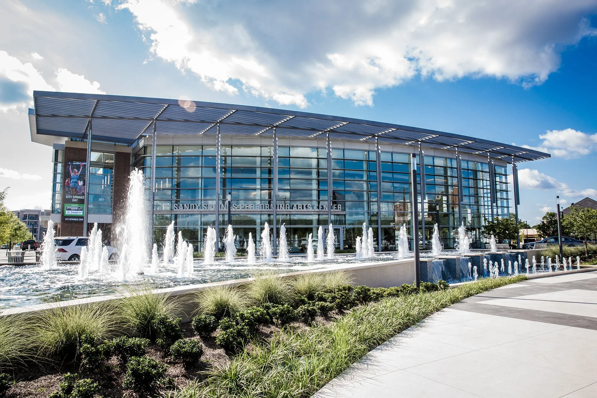 Aerial view of a residential neighborhood with multi-story apartment buildings surrounded by green trees, with a parking lot and a city skyline in the background under a partly cloudy sky.
