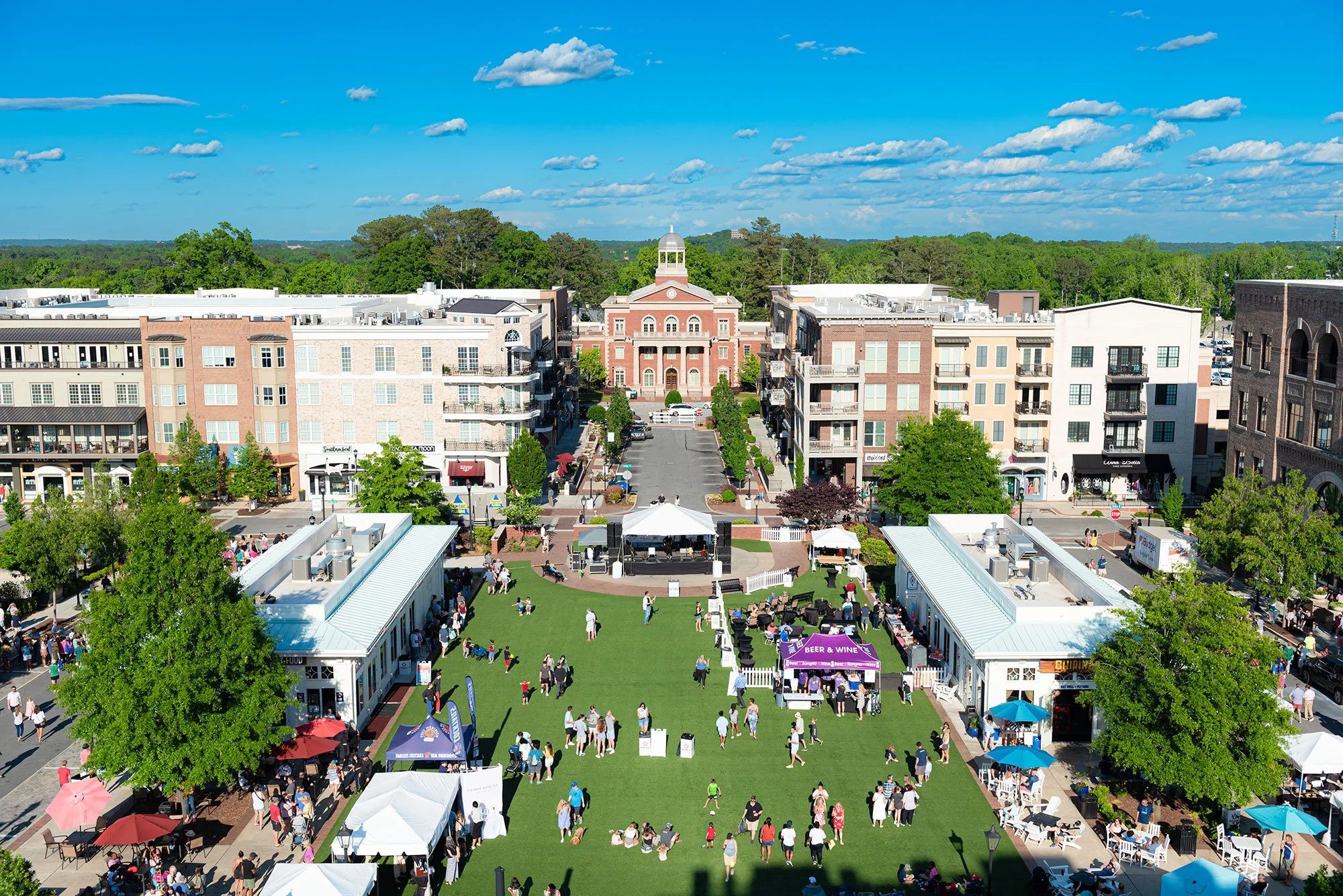 Outdoor festival in a city square with green grass, tents, and people enjoying a sunny day surrounded by modern buildings.
