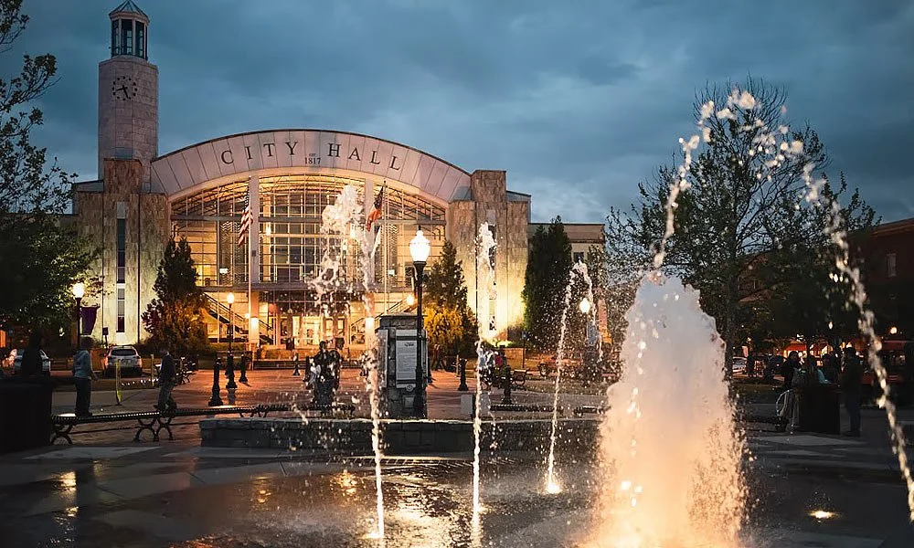 Spacious living room featuring a staircase with black railing, a large window wall with a view of an outdoor patio with umbrella, sitting area with four chairs, fireplace, and a wooden bench, with high ceilings and elegant decor.
