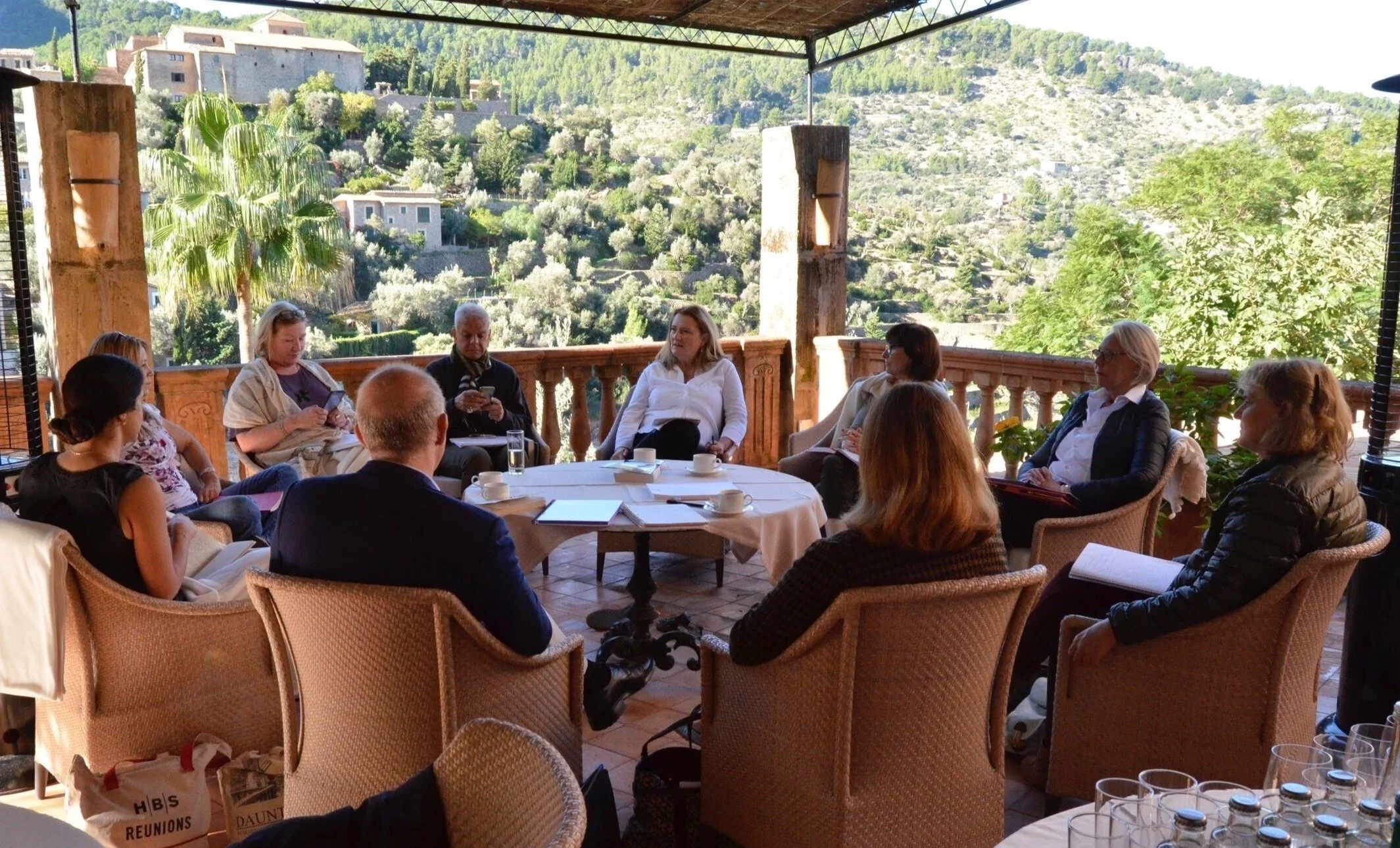 A group of people sitting around a circular table on a covered outdoor terrace, engaged in discussion, with scenic hillside landscape in the background.