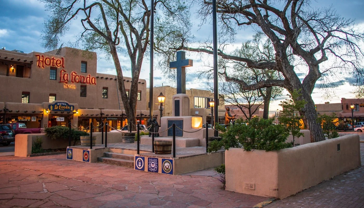 A town square with a monument featuring a cross and a statue of a person, trees, and a building with a sign reading 'Hotel La Fonda,' with a sunset sky overhead.