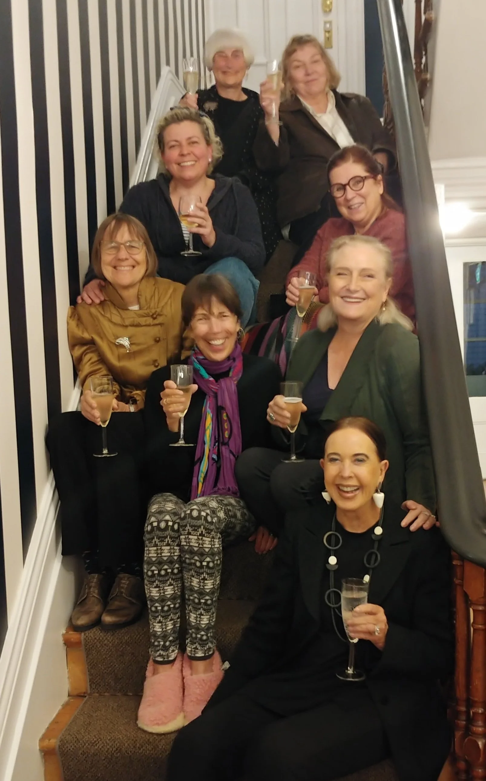 A group of nine women celebrating together on a staircase, holding glasses of champagne, smiling, and dressed in casual to semi-formal attire.