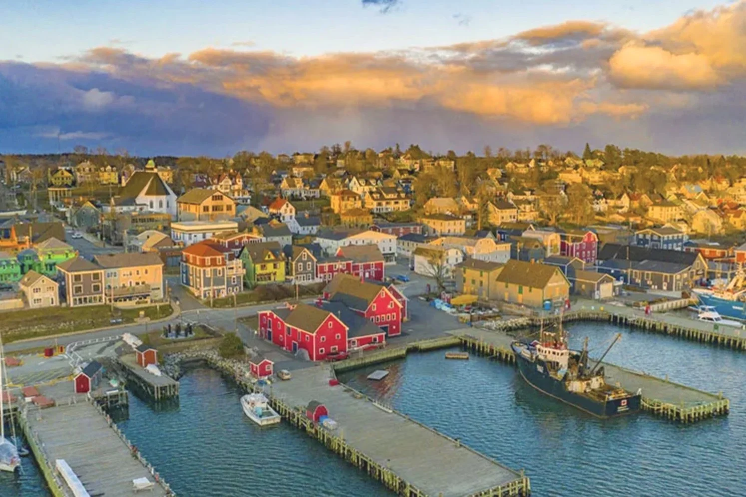 Colorful houses and buildings along a marina with boats docked, under a sky with clouds at sunset.