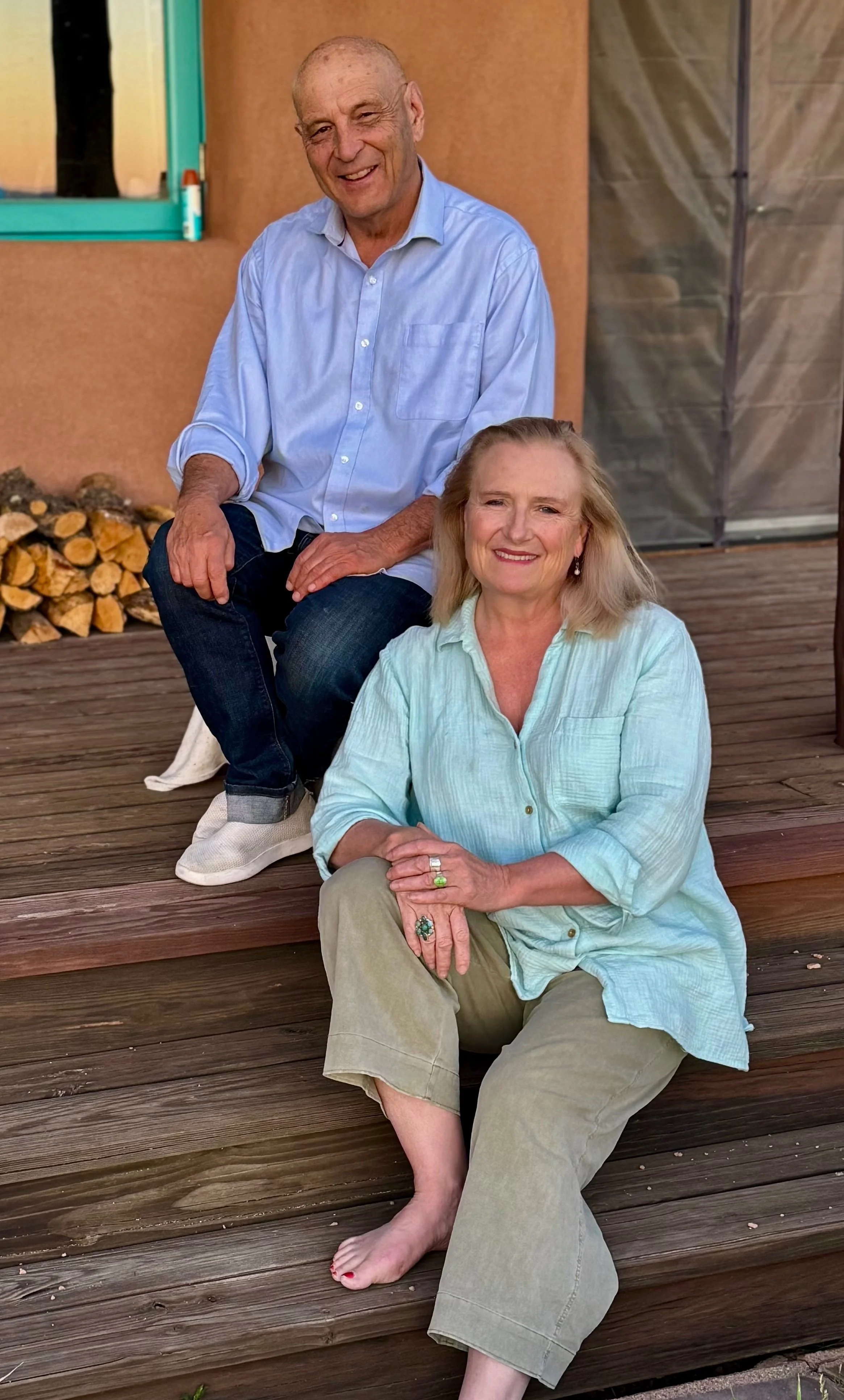 James Nave and Allegra Huston sitting on wooden steps outdoors, smiling at the camera, with a house wall, window, and stacked firewood in the background.