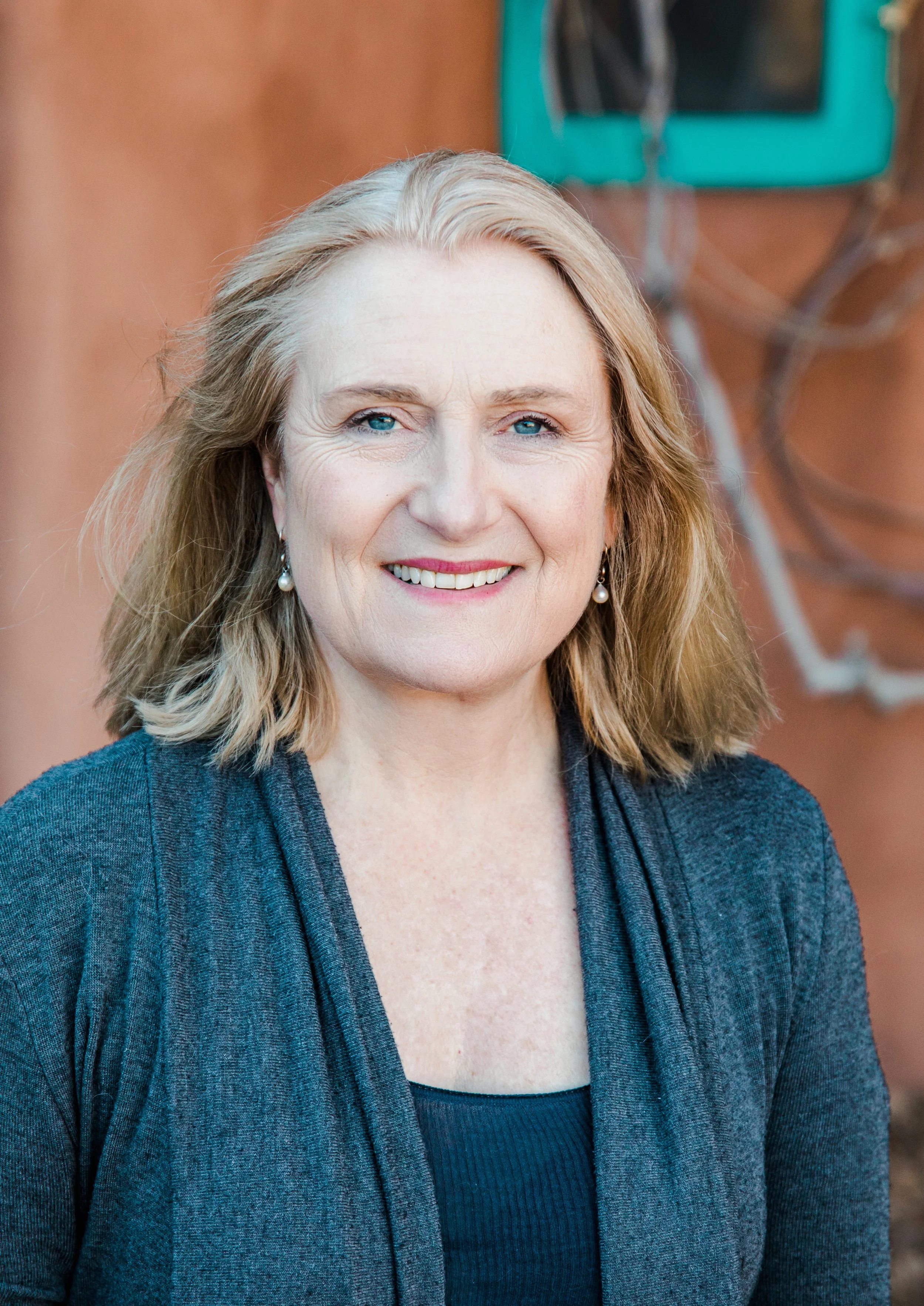 Bio photo of Allegra Huston,  with shoulder-length blonde hair, blue eyes, and earrings, smiling outdoors against a background of a Santa Fe style house.